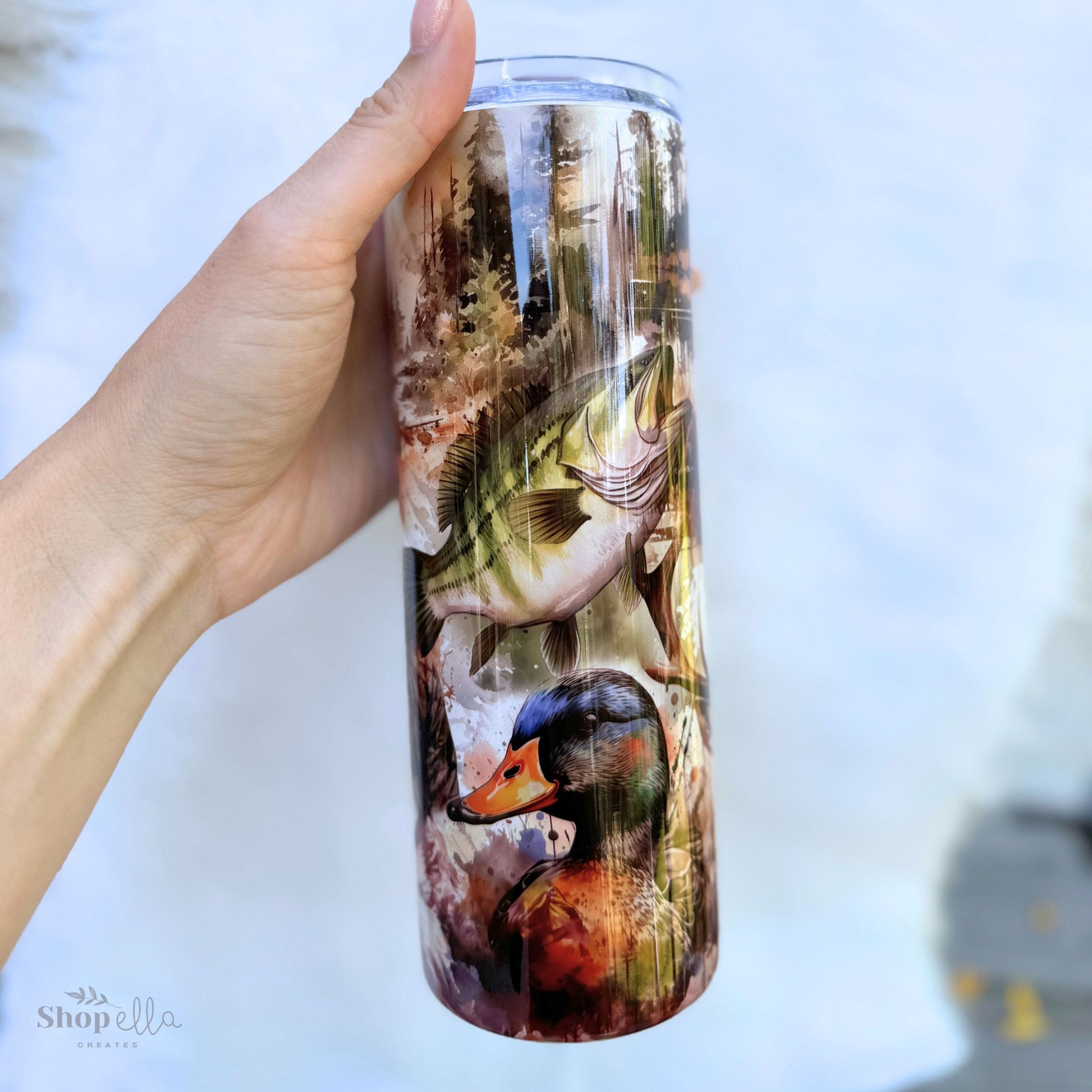 a hand holding a white 20oz skinny tumbler with lid with hunting and fishing images for men