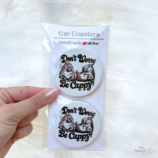 a pair of car coasters that say don't worry be cappy with a pic of two capybaras drinking margaritas and smoking. has a retro vibe